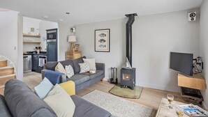 A modern living room with a sofa, a fireplace, a television, and a kitchen area visible in the background. - Churchside Cottage - 2 bed in the heart of Fowey (Fowey)