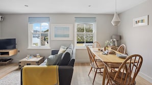 A modern living room with a sofa, a dining table with chairs, and a TV. There are windows with views of the outdoors. - Churchside Cottage - 2 bed in the heart of Fowey (Fowey)