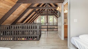 A high-ceilinged attic bedroom with exposed wooden beams, a wooden floor, and a view of trees through large windows.