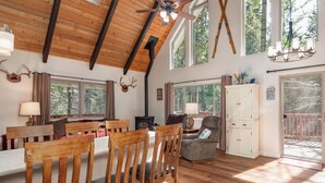 A spacious room with wooden floors, a dining table with chairs, a fireplace, and large windows offering views of the outdoors.