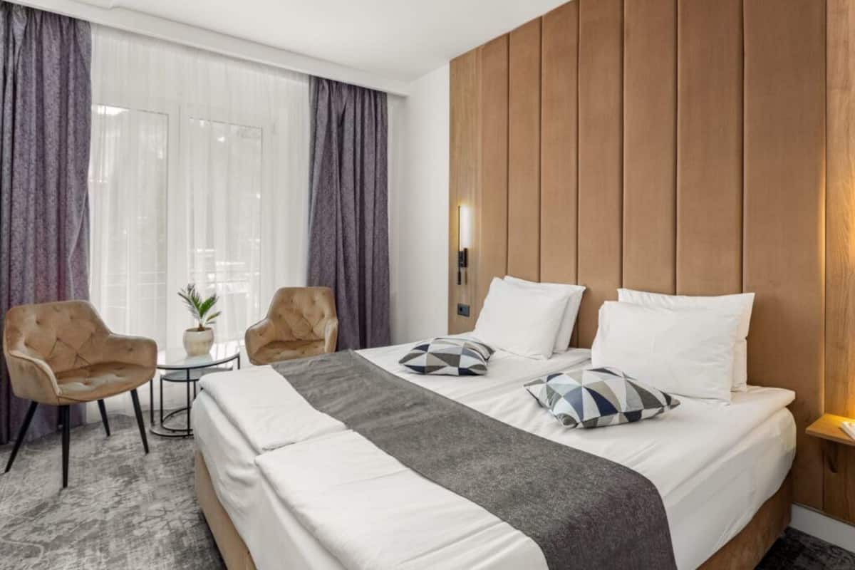 A hotel room with a large bed, two armchairs, a small table, and a potted plant.