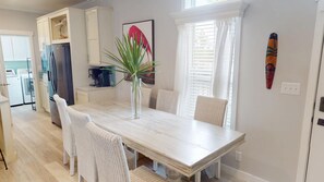 A dining area with a wooden table, white wicker chairs, a tall vase with a plant, and a kitchen in the background.