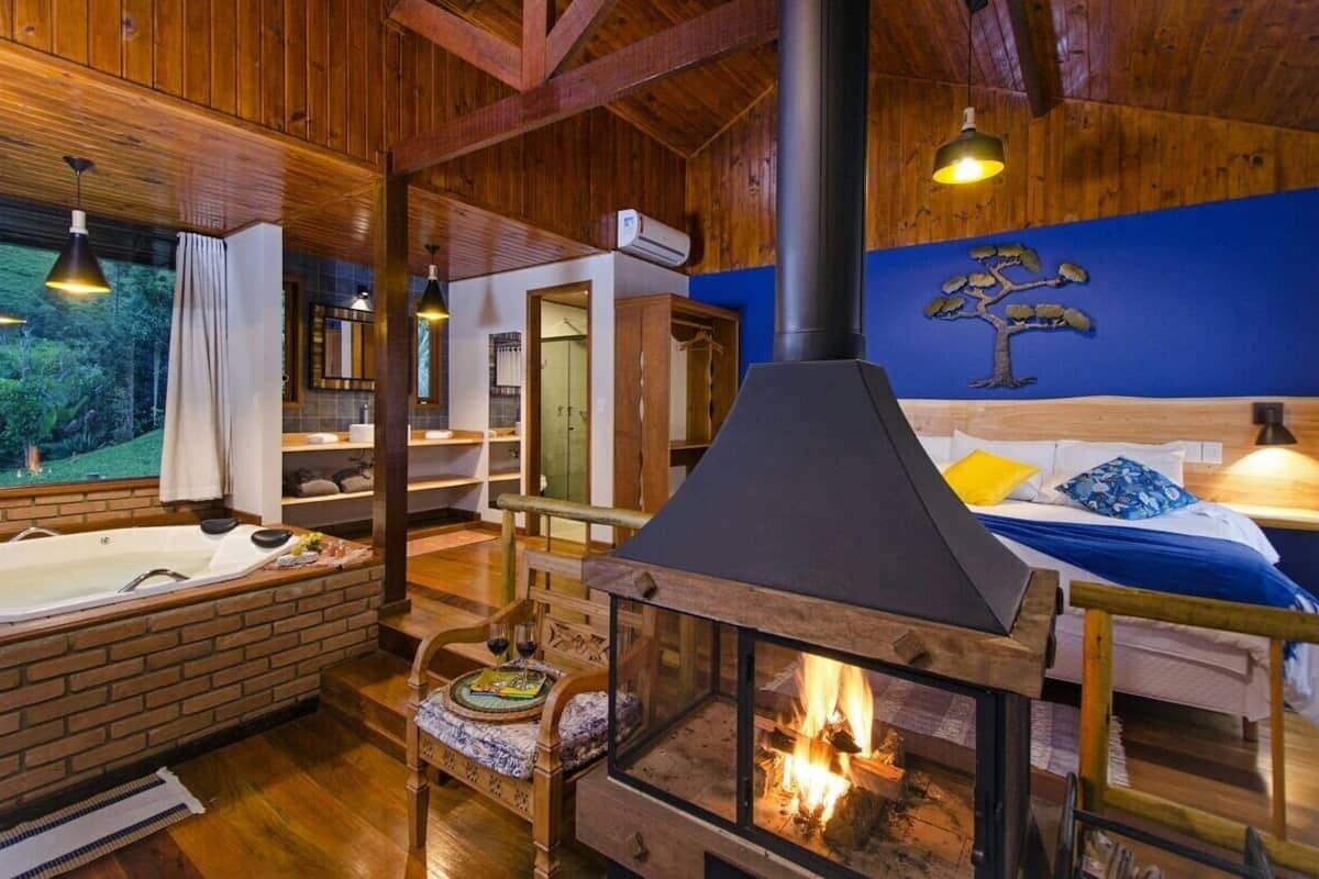 A cozy cabin interior with a wood-burning stove, a large window with curtains, a wooden bed, and a bathroom with a mirror and sink.