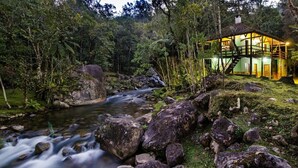 A rustic cabin with a staircase, surrounded by a lush forest and a flowing stream. - Pousada da Gruta  (Maromba)