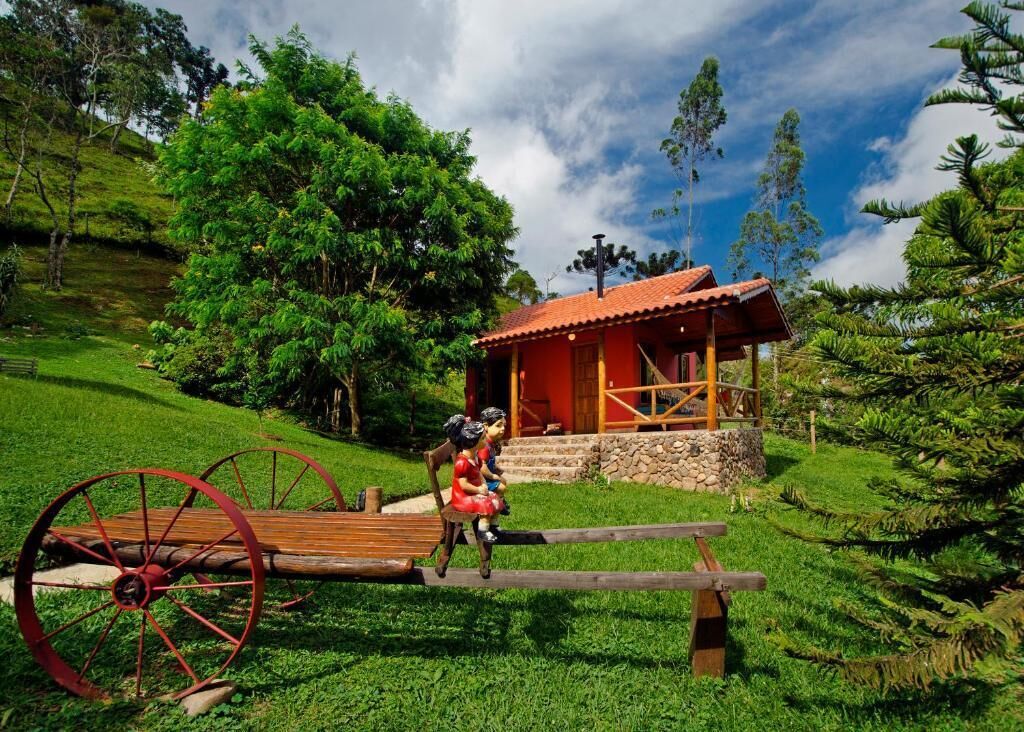 Uma carroça de madeira rústica com rodas vermelhas, um pequeno edifício com telhado vermelho e árvores verdes exuberantes ao fundo.