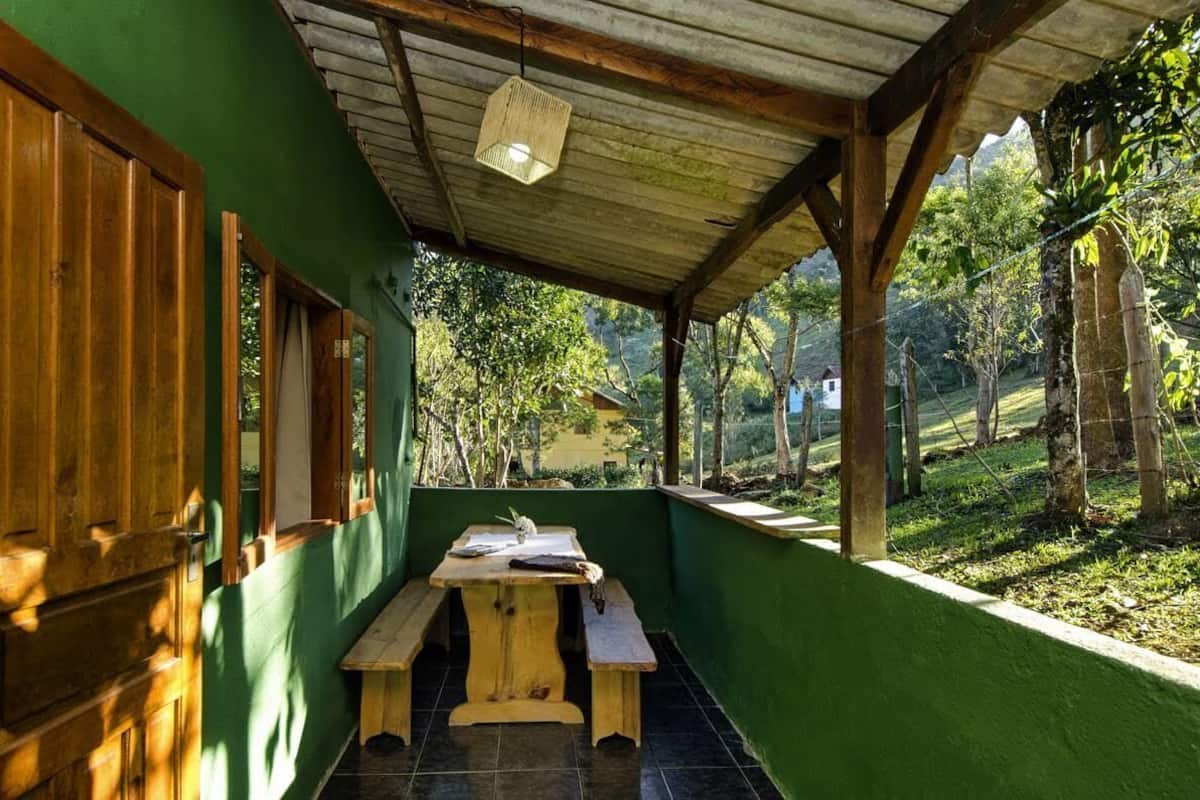 A covered outdoor area with a wooden table and benches, surrounded by greenery and a view of a hillside.