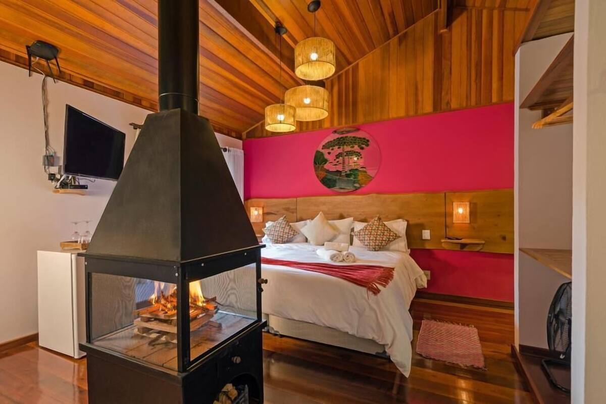 A bedroom with a bed, a wooden ceiling, a wall-mounted TV, and a fireplace.