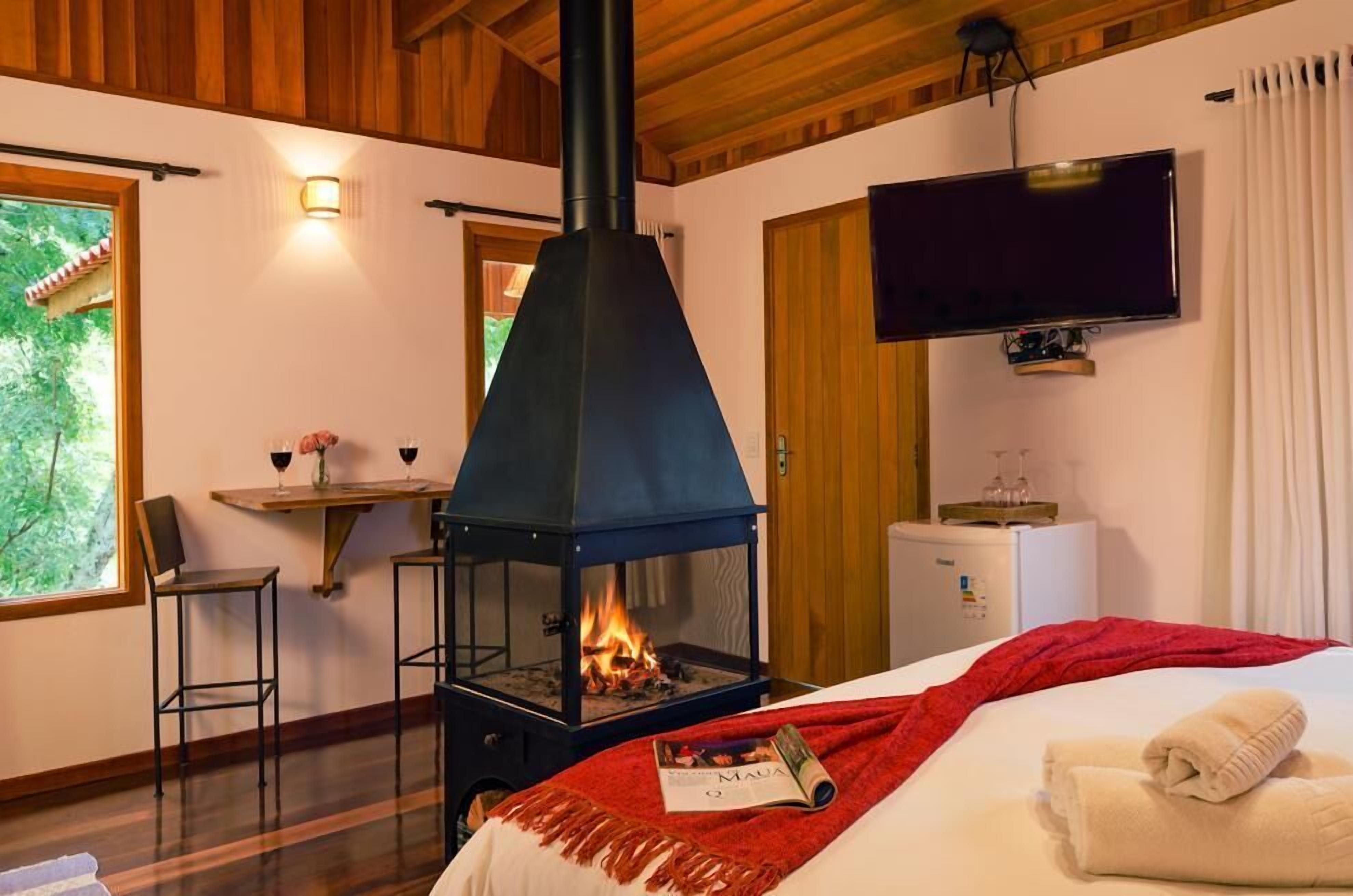 Quarto de hotel com lareira a lenha, cama com manta vermelha, televisão na parede e uma pequena mesa com uma taça de vinho.