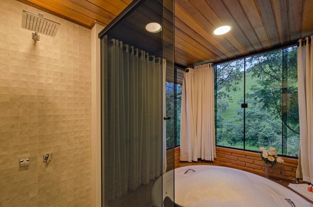 A modern bathroom with a glass shower enclosure, a large bathtub, and a view of the forest through large windows.