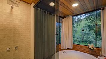 A modern bathroom with a glass shower enclosure, a large bathtub, and a view of the forest through large windows.