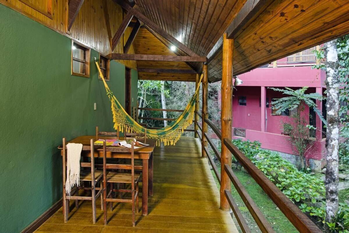 A wooden veranda with a hammock, a table with chairs, and a view of a red building and greenery.