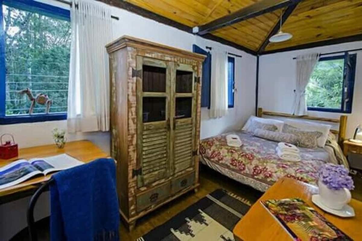 A cozy room with a wooden ceiling, a bed with floral bedding, a wooden wardrobe, a desk with a chair, and a window with a view of trees.