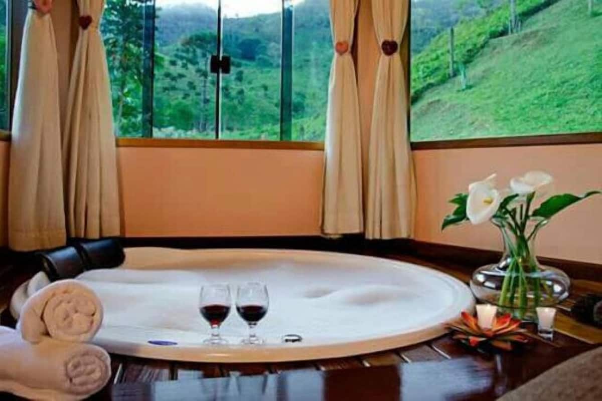 A hotel room with a large window, a round dining table with two wine glasses, a vase with flowers, and a rolled towel on the floor.