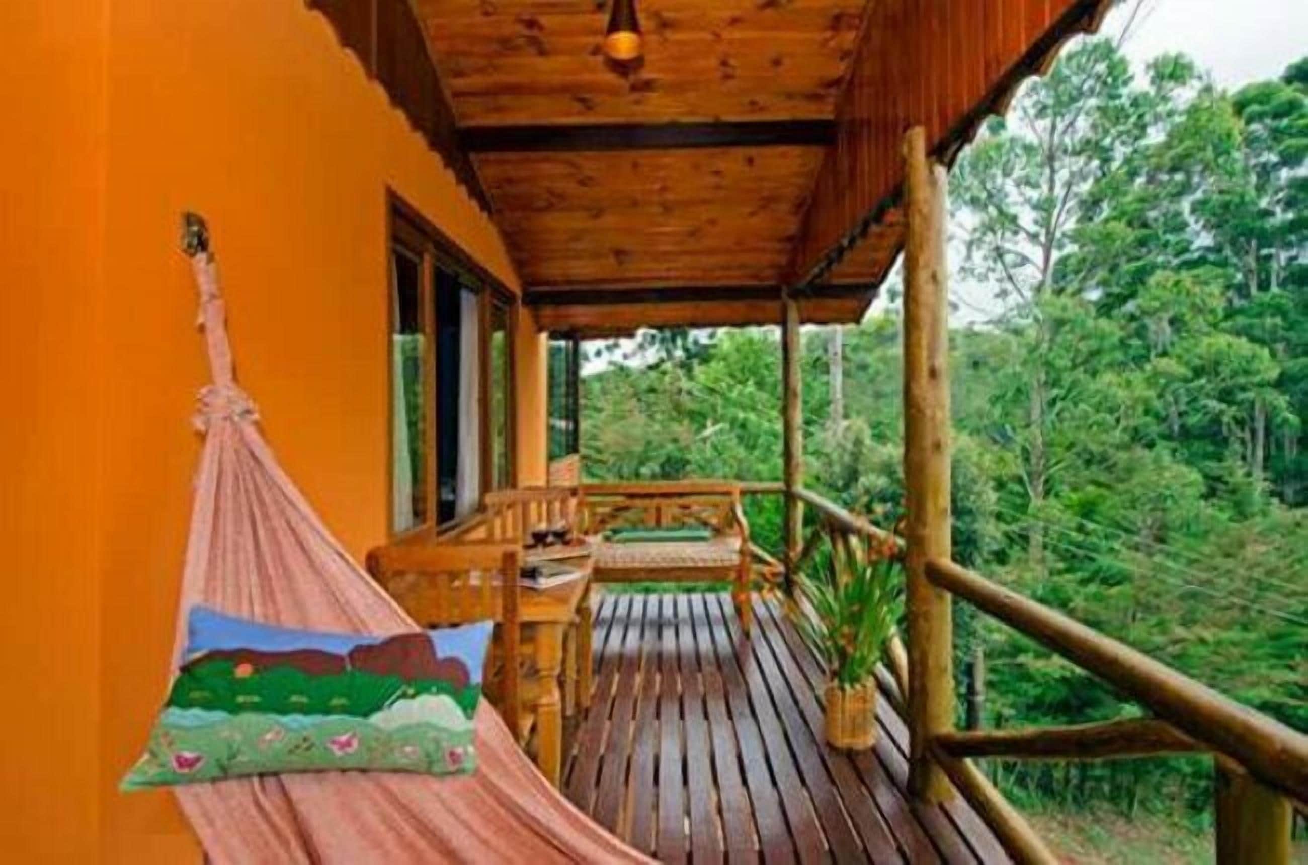 Una terraza de madera con una hamaca, una mesa y sillas, con vista a una zona boscosa.