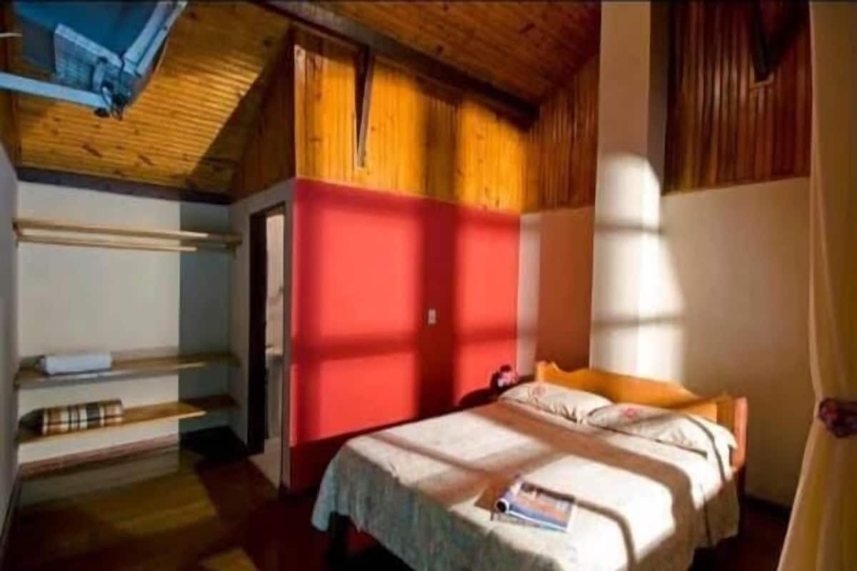 A bedroom with a wooden ceiling, a red wall, a bed with a white bedspread, and a window with curtains.