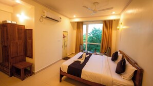 A hotel room with a bed, a ceiling fan, an air conditioning unit, a wooden wardrobe, a small table, and a window with curtains. - Hotel Marine (Varanasi)