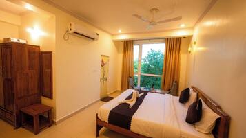 A hotel room with a bed, a ceiling fan, an air conditioning unit, a wooden wardrobe, a small table, and a window with curtains.