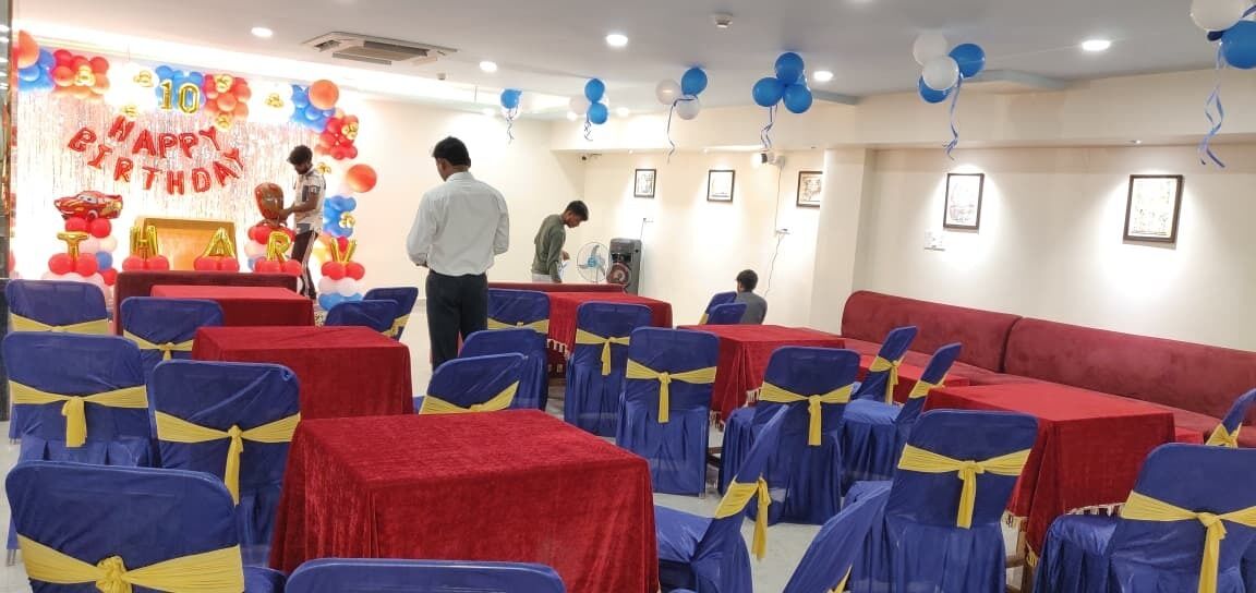Une salle avec des rangées de tables et de chaises, dont certaines sont recouvertes de nappes rouges, et une scène décorée de ballons et d’une banderole.
