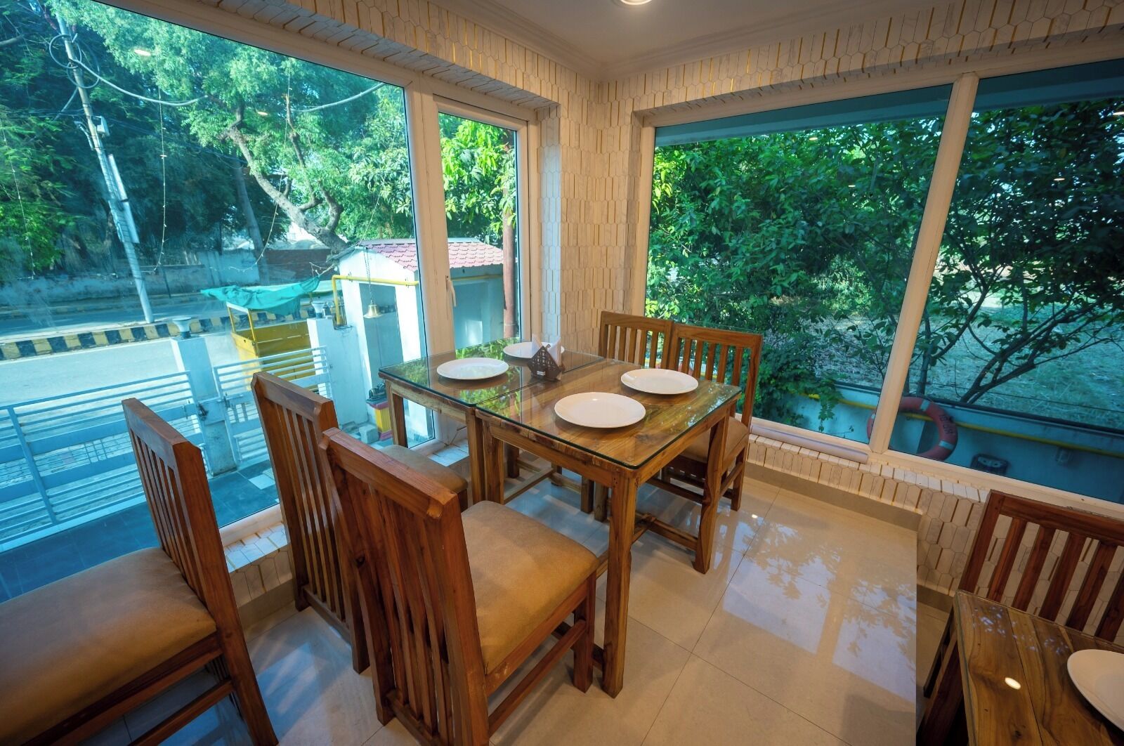 A dining area with a glass table and wooden chairs, large windows with a view of trees and a road, and a tiled floor.