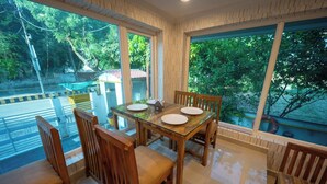 A dining area with a glass table and wooden chairs, large windows with a view of trees and a road, and a tiled floor. - Hotel Marine (Varanasi)