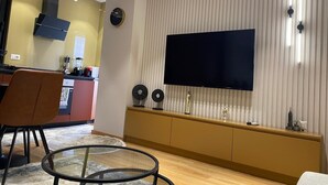 A modern living room with a flat-screen TV mounted on a wall, a glass coffee table, and a seating area with a chair. - Florenc's Apartment (Farke)