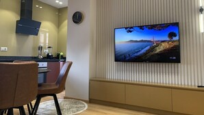 A modern living room with a flat-screen TV mounted on a wall, a dining area with a table and chairs, and a kitchen with a built-in stove and extractor hood. - Florenc's Apartment (Farke)
