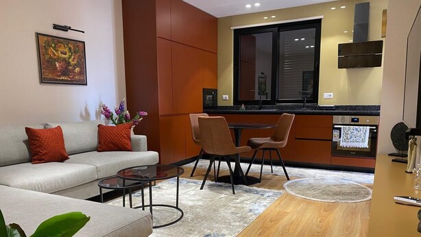 A modern living room with a sofa, coffee table, and chairs. There is a kitchen area with a window and cabinets. - Florenc's Apartment (Farke)