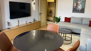 A modern living room with a flat-screen TV mounted on a wall, a round glass table, and a grey sofa with red cushions. - Florenc's Apartment (Farke)