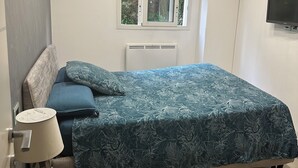 A bedroom with a bed, a nightstand, a lamp, a window, and a TV.