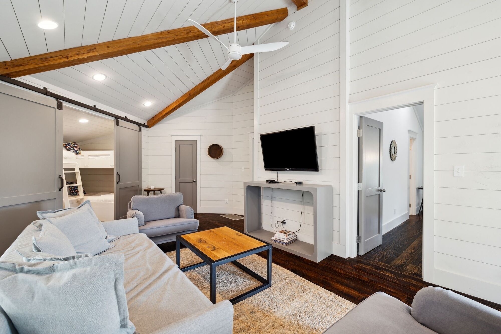 Un salon moderne doté d’un plafond haut, de poutres apparentes en bois, d’un téléviseur à écran plat fixé à un mur blanc, d’un canapé gris, d’une table basse en bois et d’une cheminée.