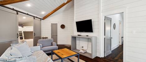 Un salon moderne doté d’un plafond haut, de poutres apparentes en bois, d’un téléviseur à écran plat fixé à un mur blanc, d’un canapé gris, d’une table basse en bois et d’une cheminée.