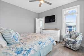 A bedroom with a bed, a dresser, a chair, a ceiling fan, and a TV on the wall.