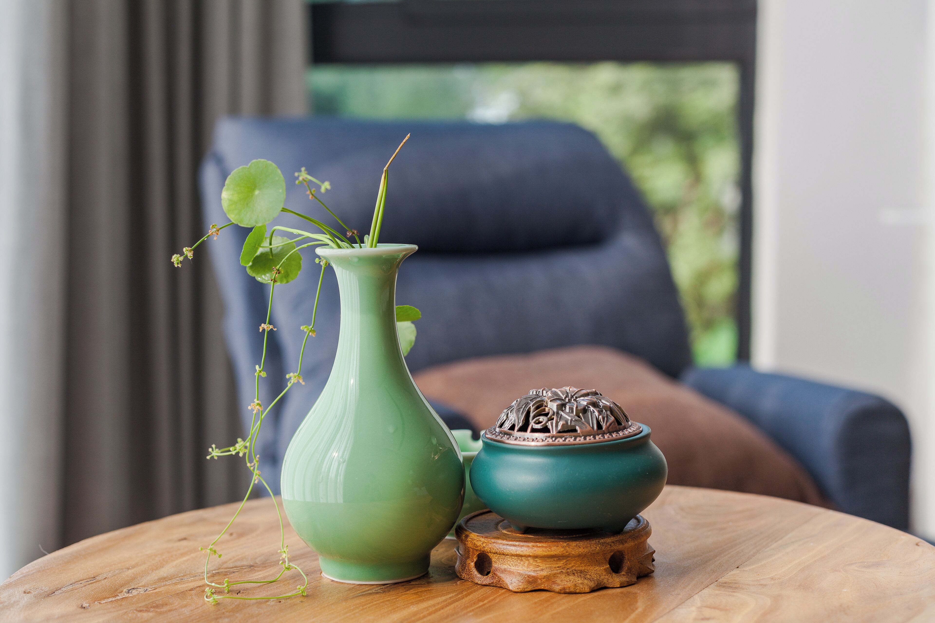 A green vase with a plant on a wooden table, a small green pot, and a blue chair in the background.