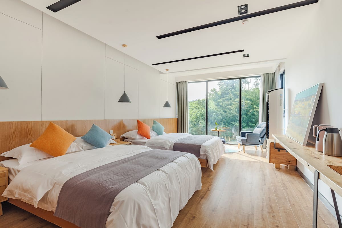 Ein Hotelzimmer mit zwei Betten, einem Schreibtisch mit Wasserkocher und einem großen Fenster mit Blick auf Bäume.