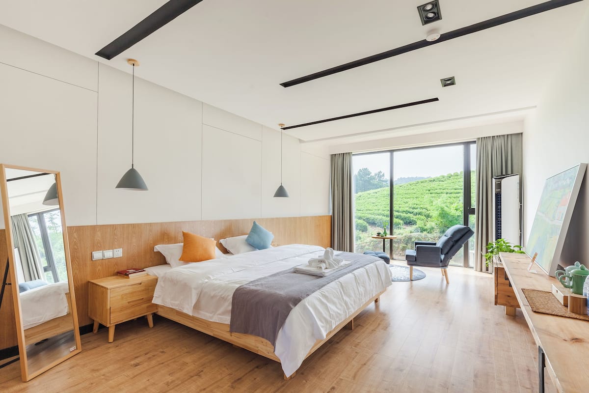 Ein modernes Hotelzimmer mit einem großen Bett, Nachttischen, einem Schreibtisch mit Stuhl, einem Spiegel und Blick auf Grünfläche.
