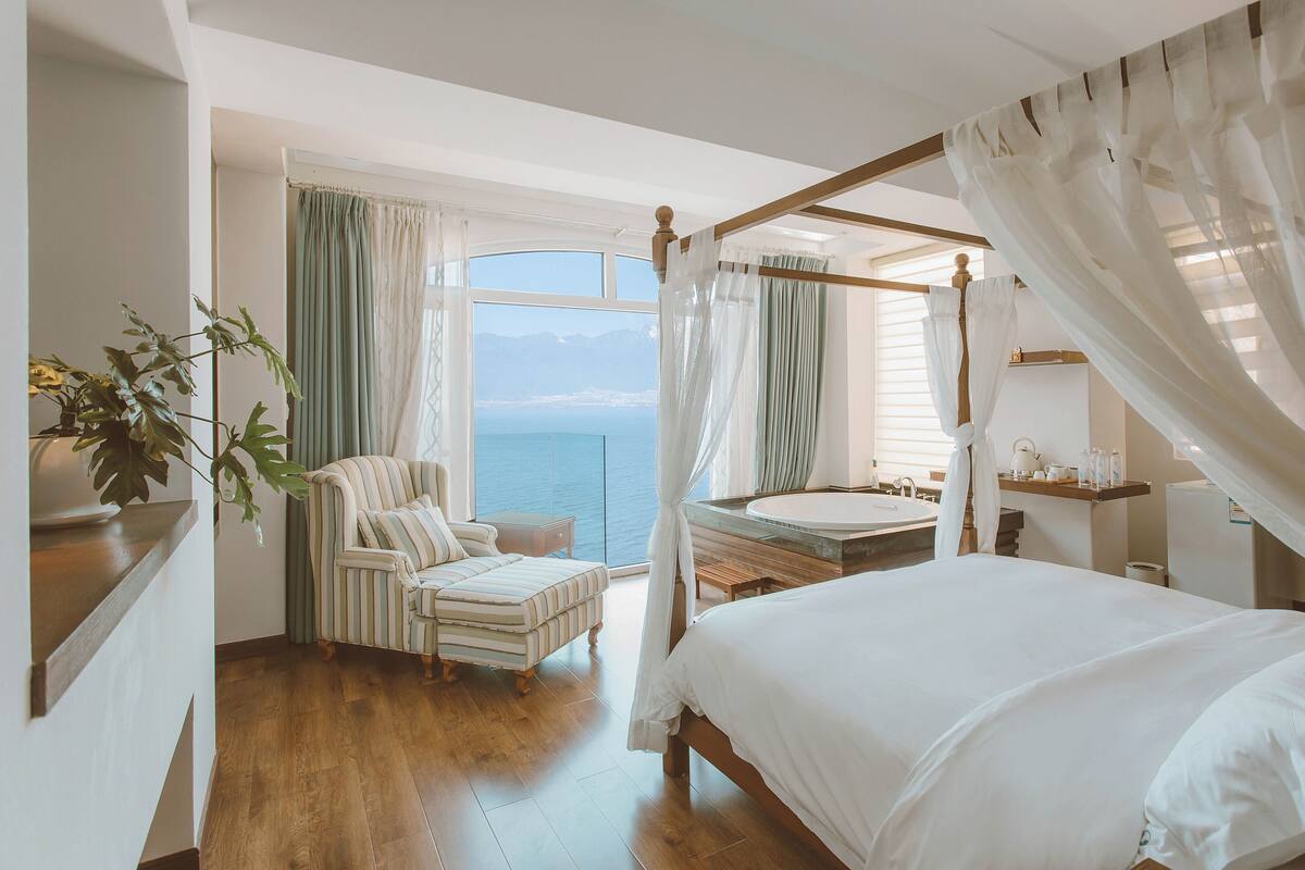 Une chambre à coucher avec un lit à baldaquin, un coin salon comprenant un fauteuil et une petite table, et une vue sur la mer.