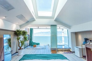 A modern hotel room with a large window offering a view of the sea, a bed, a seating area with a view, and a small kitchenette. - Dali Mermaid Sea View Resort Hotel (Dali)