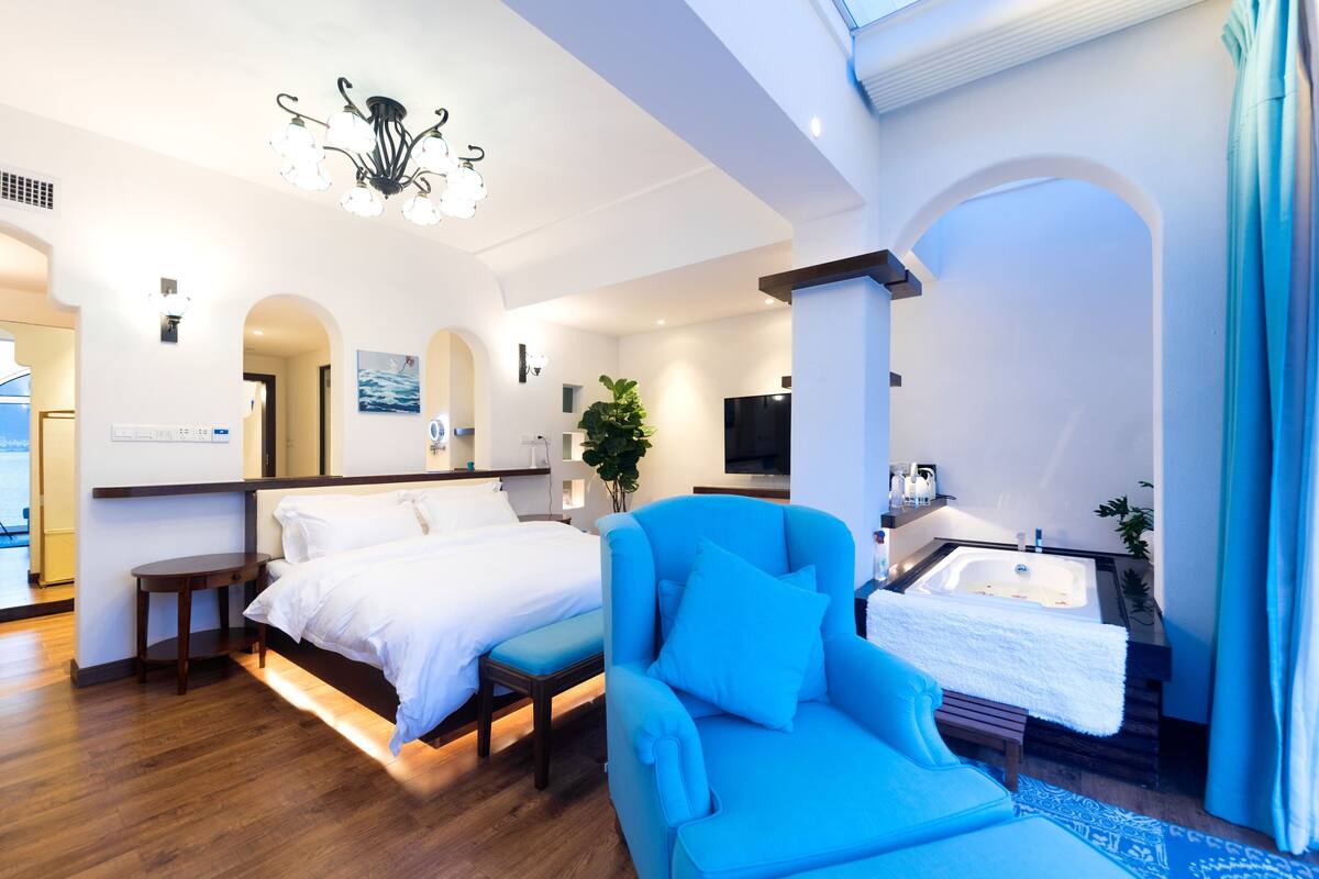 Une chambre d’hôtel moderne avec un grand lit, un fauteuil bleu, une baignoire et un lustre.