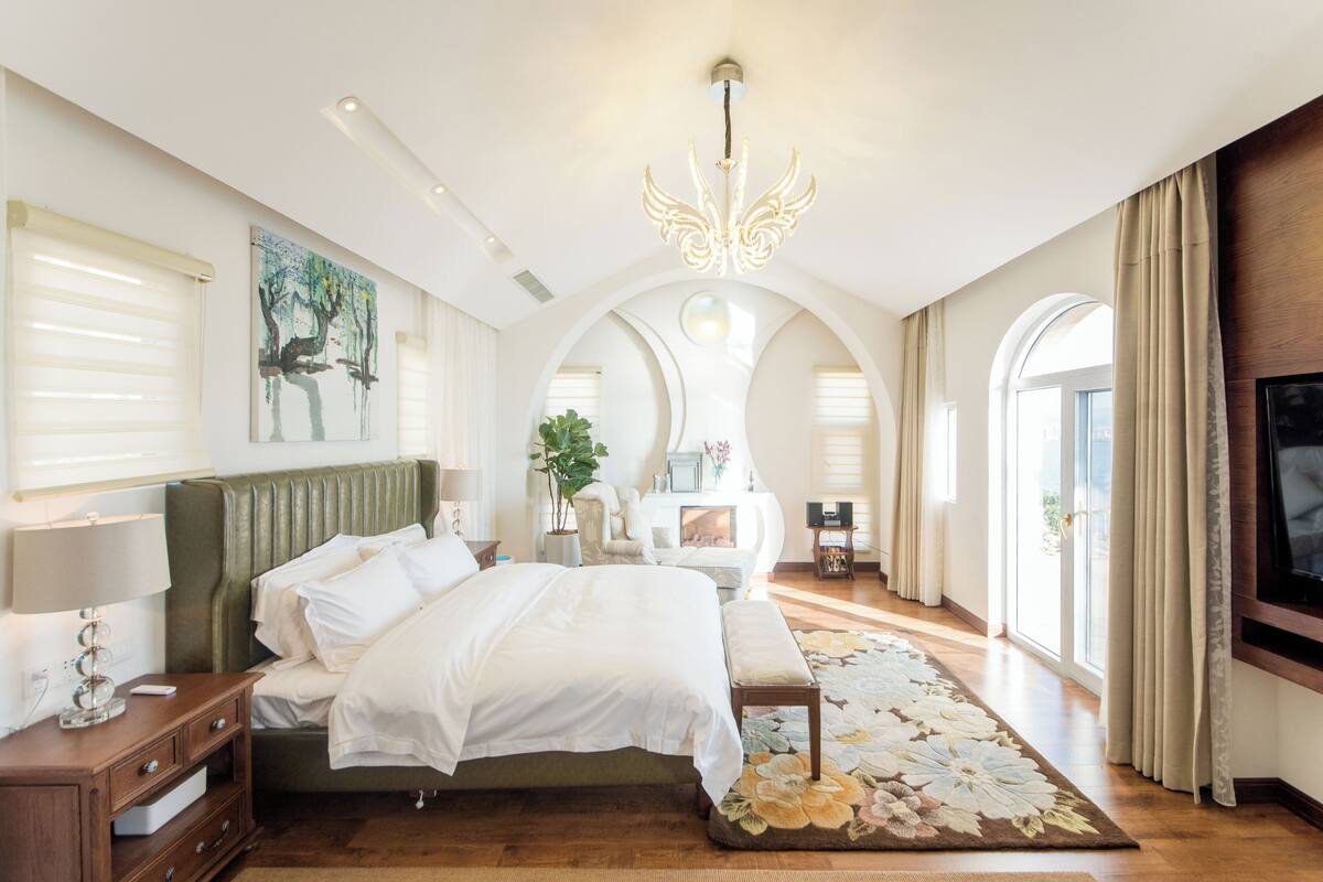 Une chambre spacieuse avec un grand lit, un lustre, un tableau au mur et un téléviseur fixé au mur.