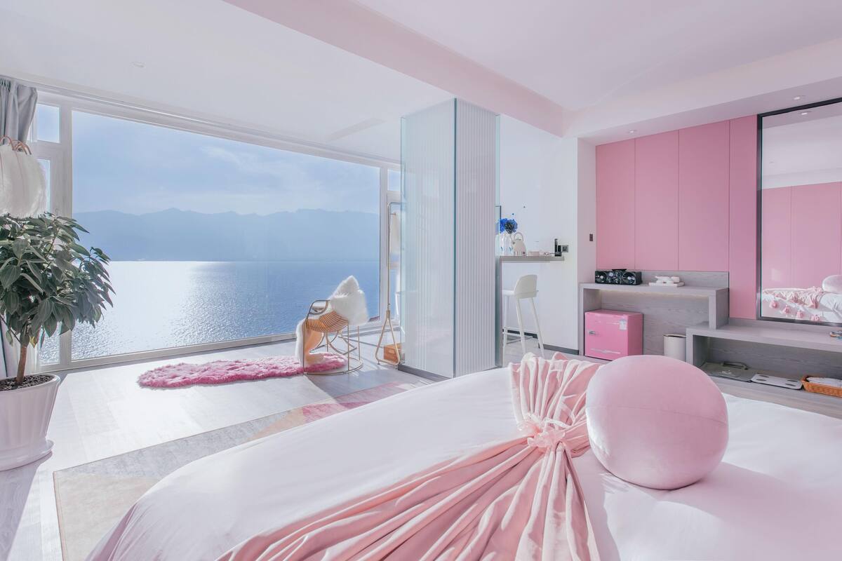 Une chambre moderne avec une grande fenêtre donnant sur un plan d’eau et des montagnes, un lit avec une literie rose, une chaise blanche et une commode rose.