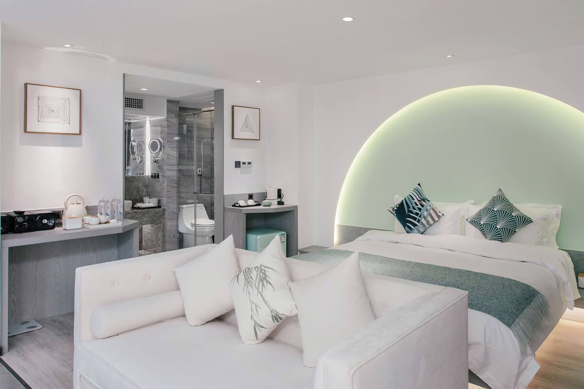 Une chambre moderne avec un plafond incurvé, un lit agrémenté d’oreillers à motifs, un canapé blanc et une salle de bain dotée d’une cabine de douche en verre.