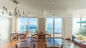 A dining area with a large wooden table and chairs, large windows with a view of the sea, and a modern chandelier.