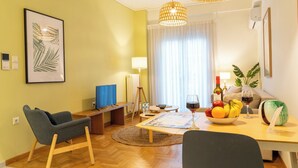 A cozy living room with a wooden floor, a blue sofa, a dining table with fruit and wine, a lamp, and a framed plant. - Acropolis Central Living (Athina)