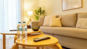 A living room with a sofa, a coffee table with water bottles and a remote control, a lamp, and a plant. - Acropolis Central Living (Athina)