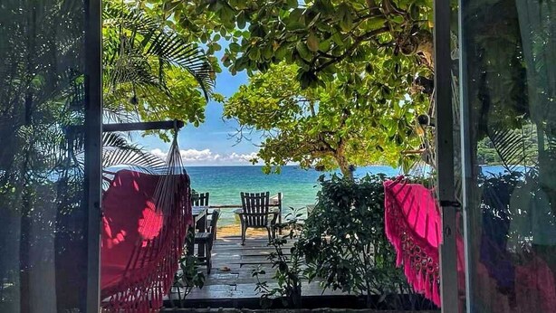 A view from inside a room to a wooden deck with a table and chairs, overlooking a beach. - Hostal Hilltop Sapzurro (Sapzurro)
