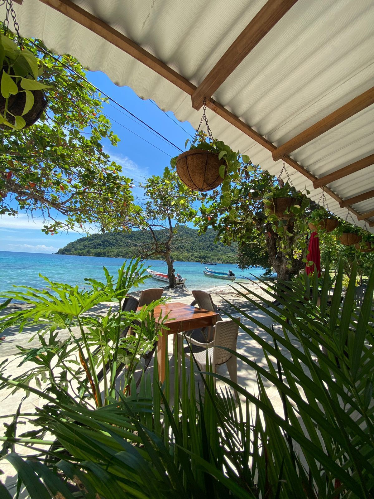 Un área de estar exterior techada con una mesa y sillas, rodeada de plantas colgantes y vistas a una playa con barcos.