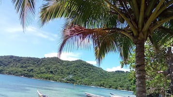 Paisaje costero con barcos en aguas tranquilas, cielo despejado y una verde colina al fondo.
