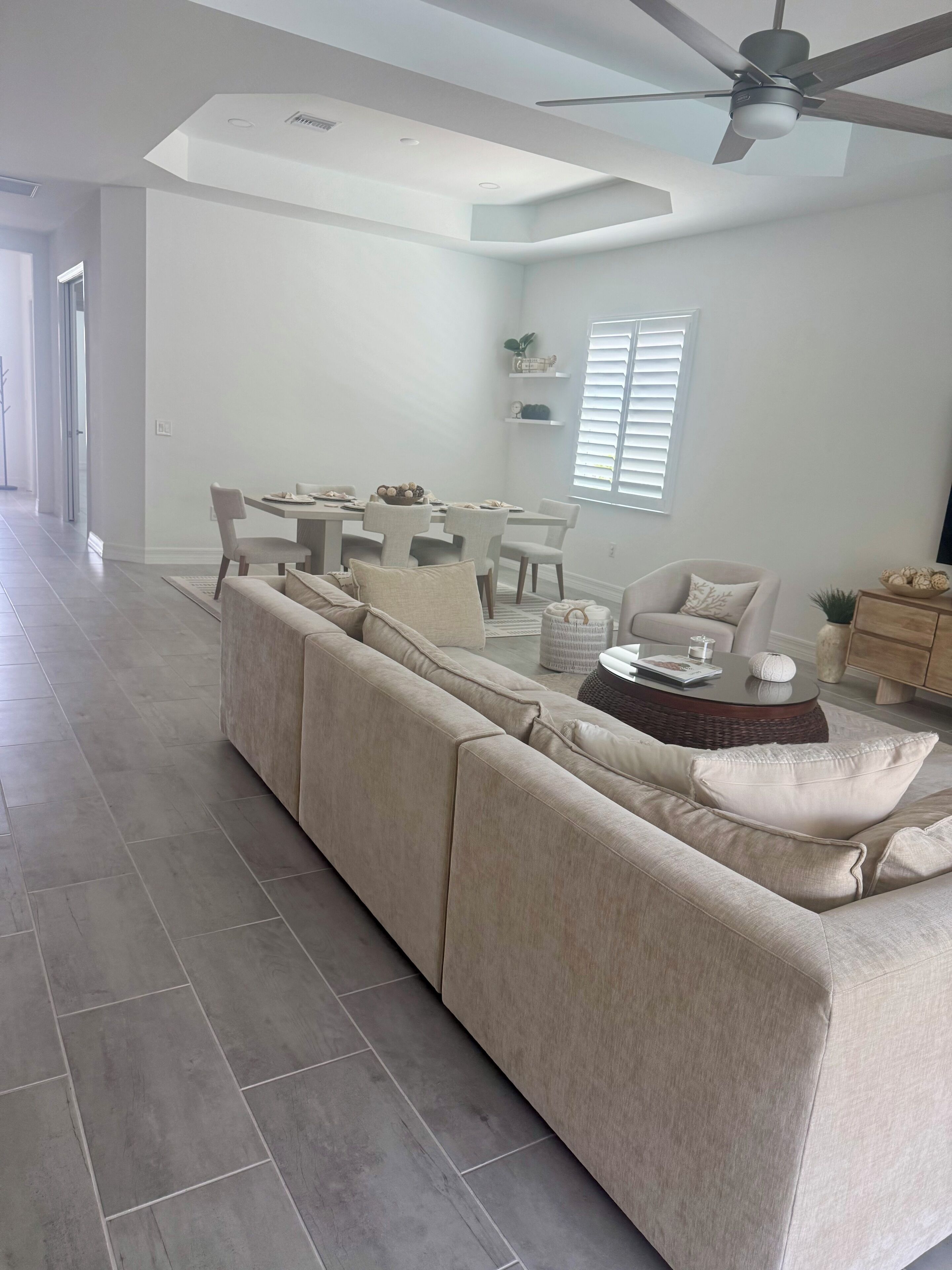 A spacious living room with a large beige sectional sofa, a dining area with a white table and chairs, and a ceiling fan.