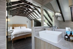 A modern bedroom with a large bed, a glass-enclosed bathroom with a freestanding bathtub, and a large mirror reflecting the room.