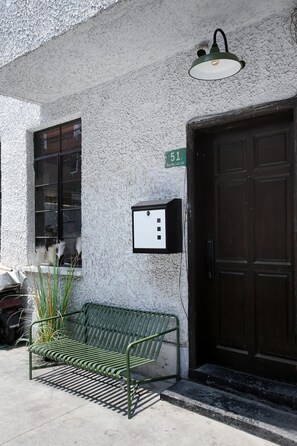 A green metal bench outside a building with a white textured wall, a black door, and a green sign with the number 51.